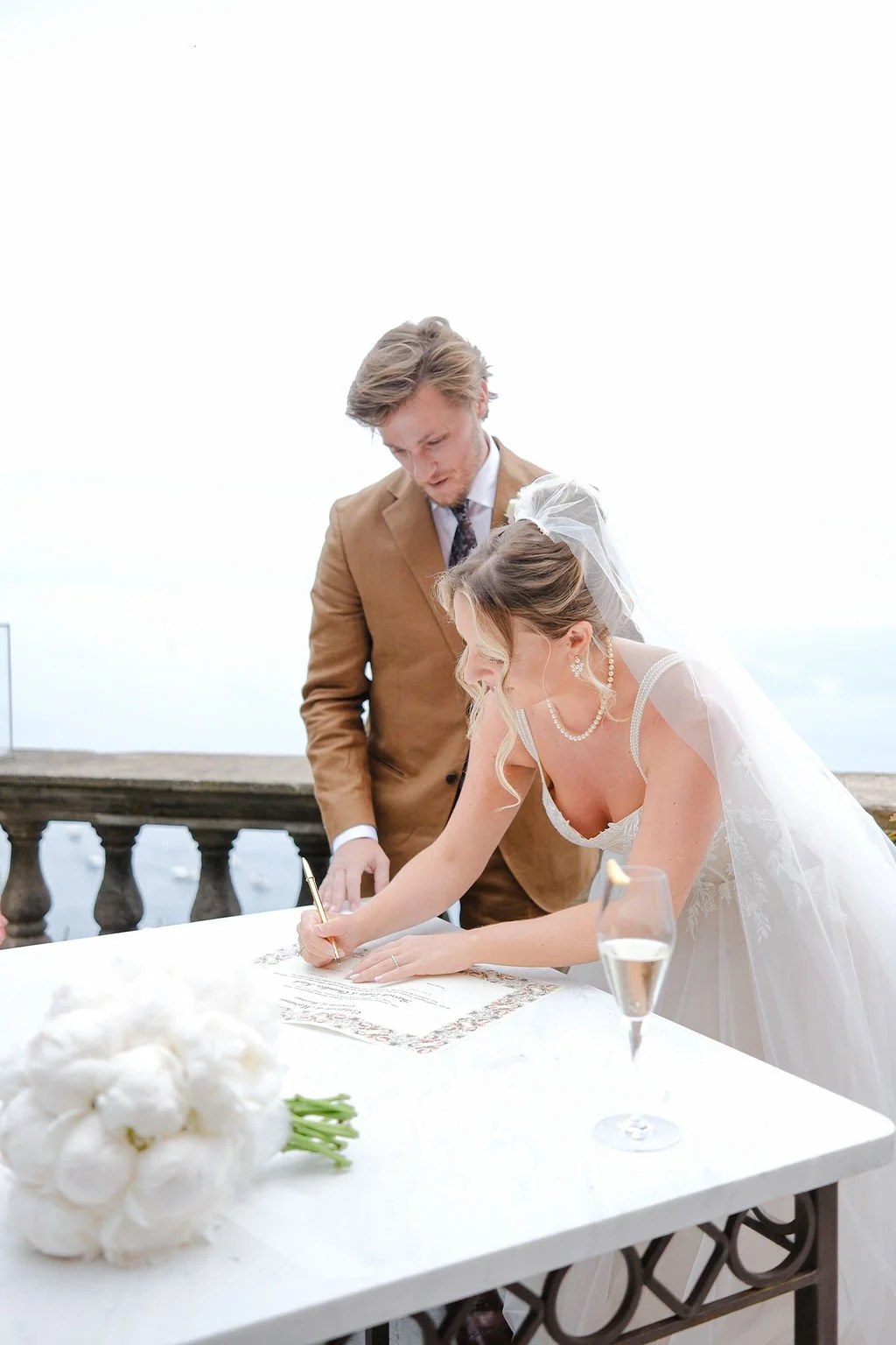 A bride signing a marriage certificate at an outdoor wedding ceremony with a groom watching, on a balcony overlooking the water, with a bouquet of white flowers and a glass of champagne on the table.