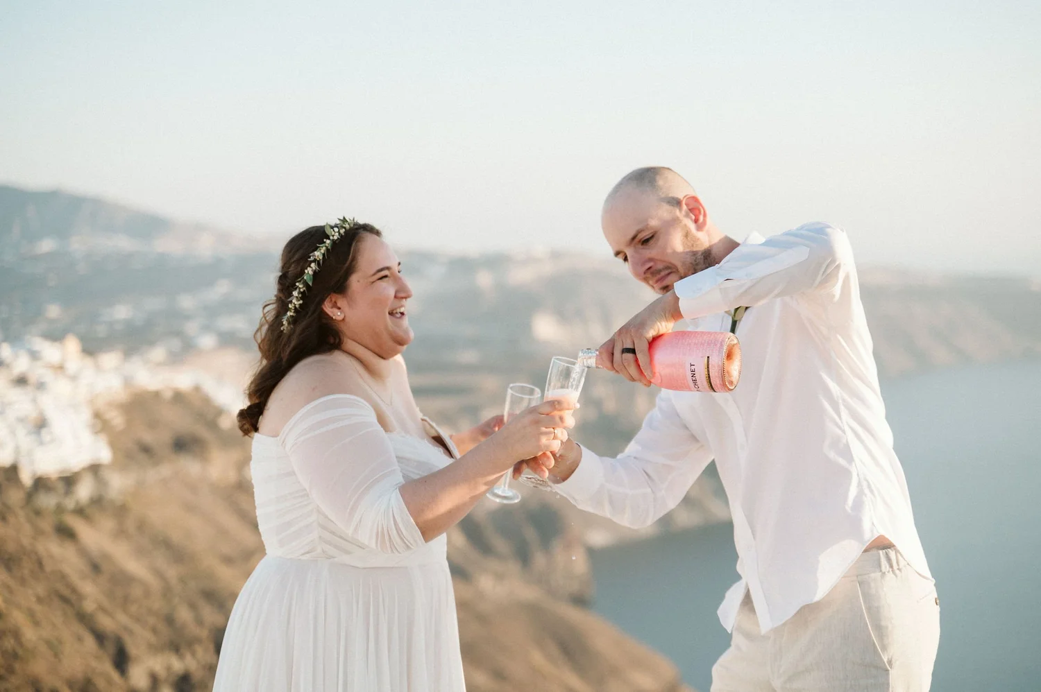 A couple celebrating at a scenic outdoor location, with the man pouring champagne into the woman's glass. The woman is wearing a white dress and a floral headband, with a backdrop of cliffs and the ocean.