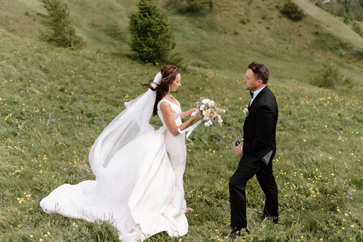 A bride in a white wedding dress holding a bouquet and a groom in a black tuxedo standing in a green field with small yellow and purple flowers, surrounded by trees and rolling hills.