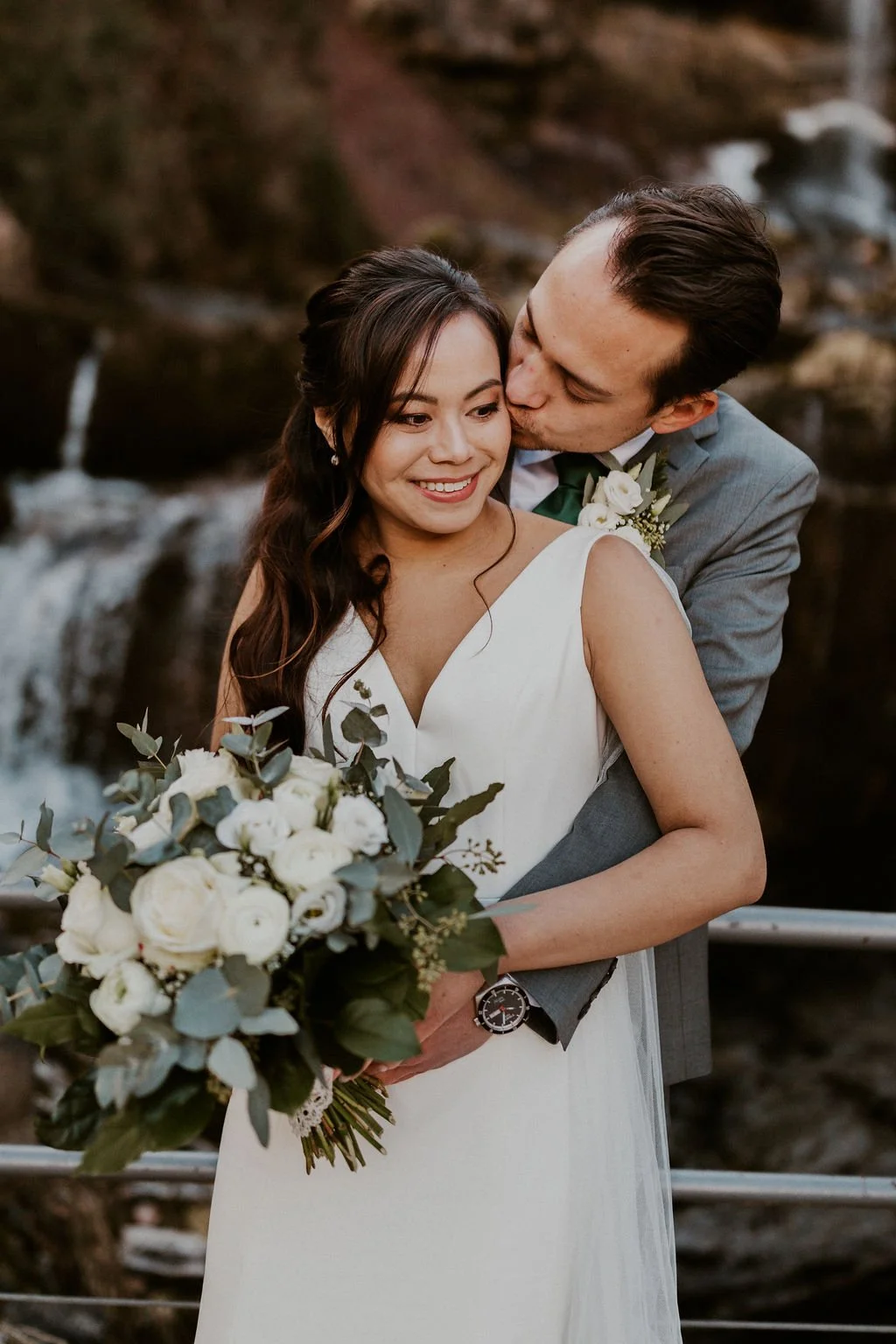 A wedding couple, woman in a white dress holding a bouquet, man in a gray suit, kissing her on the cheek, outdoors near waterfalls.