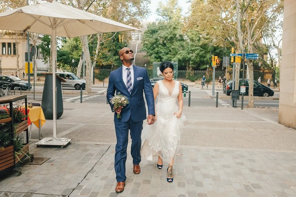 A newlywed couple walking outdoors on a city street, the groom holding a bouquet and dressed in a blue suit with a tie, the bride in a white lace wedding dress with high heels, under an umbrella, with trees and cars in the background.