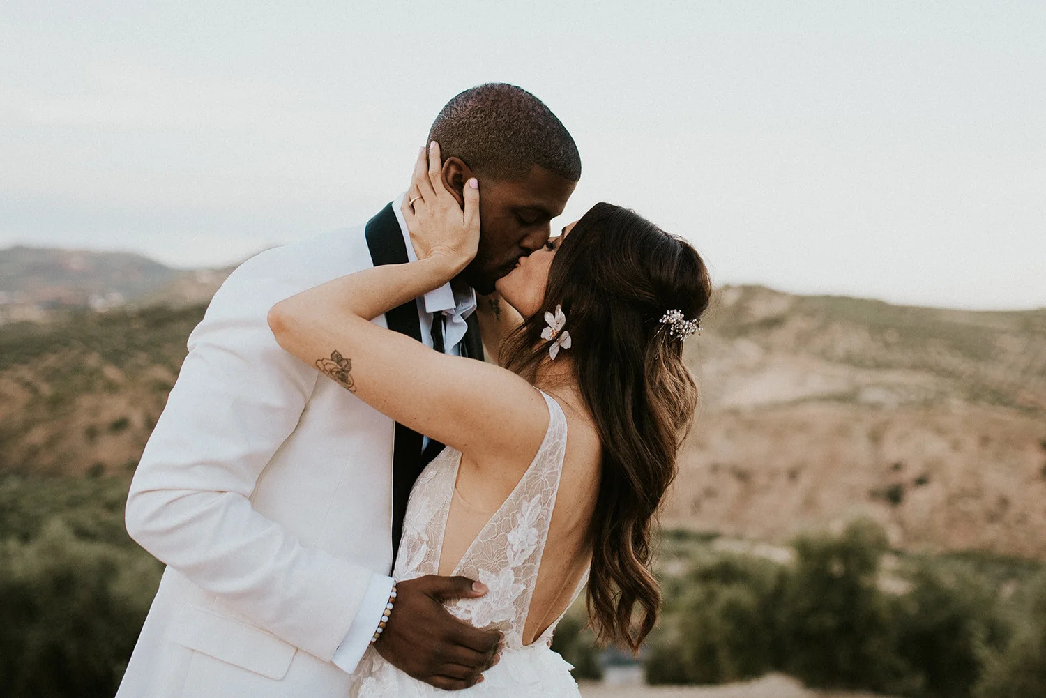 A couple in wedding attire sharing a kiss outdoors, with hilly landscape in the background.
