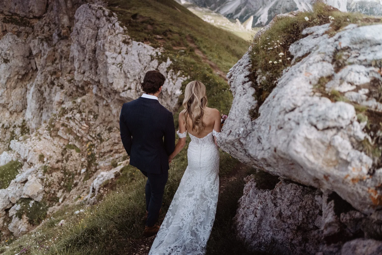 A bride and groom standing outdoors among rocky terrain and green hills, viewed from behind, with the bride in a white lace wedding dress and the groom in a dark suit.