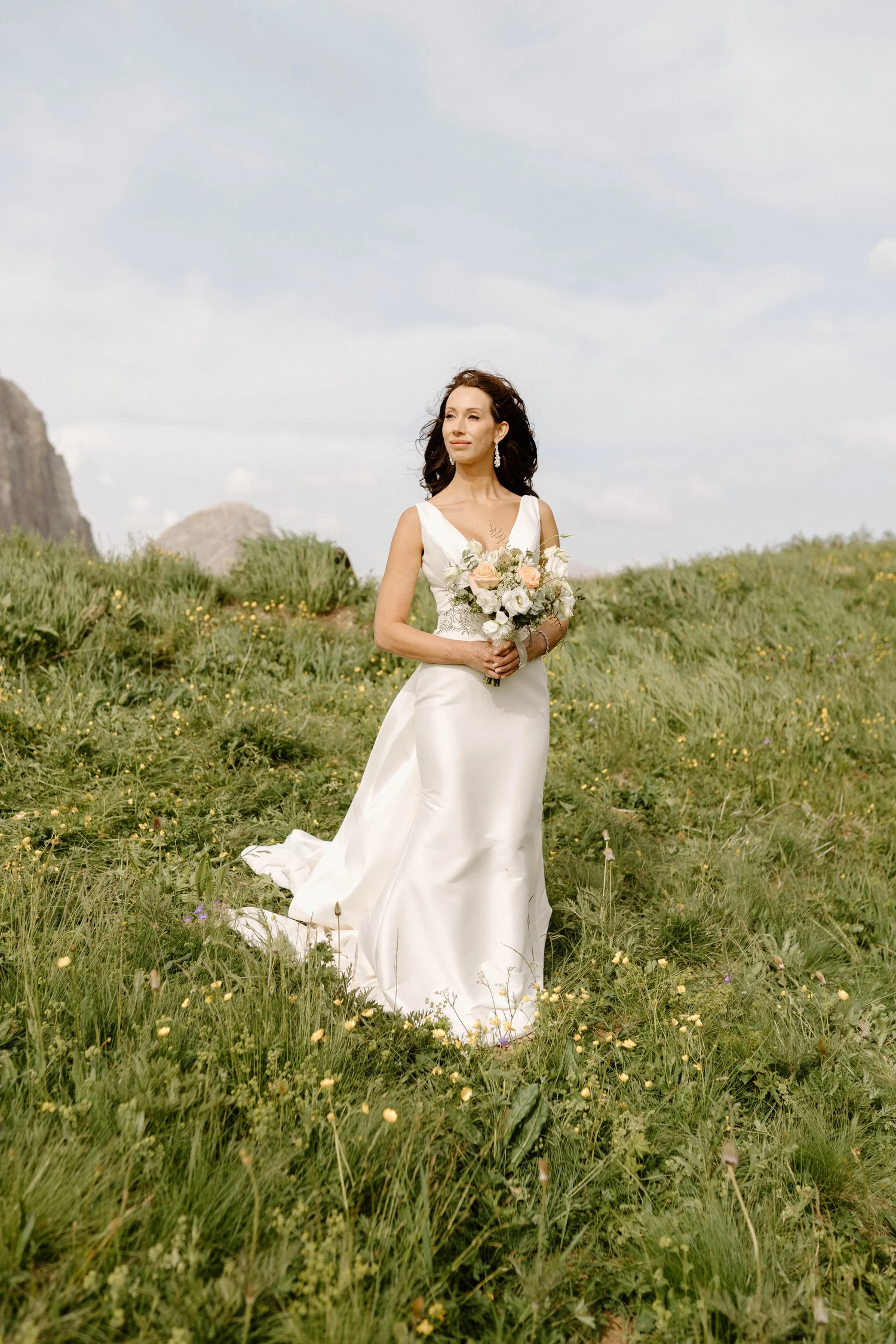 A woman in a white wedding dress standing in a grassy field holding a bouquet of flowers, with mountains and a cloudy sky in the background.