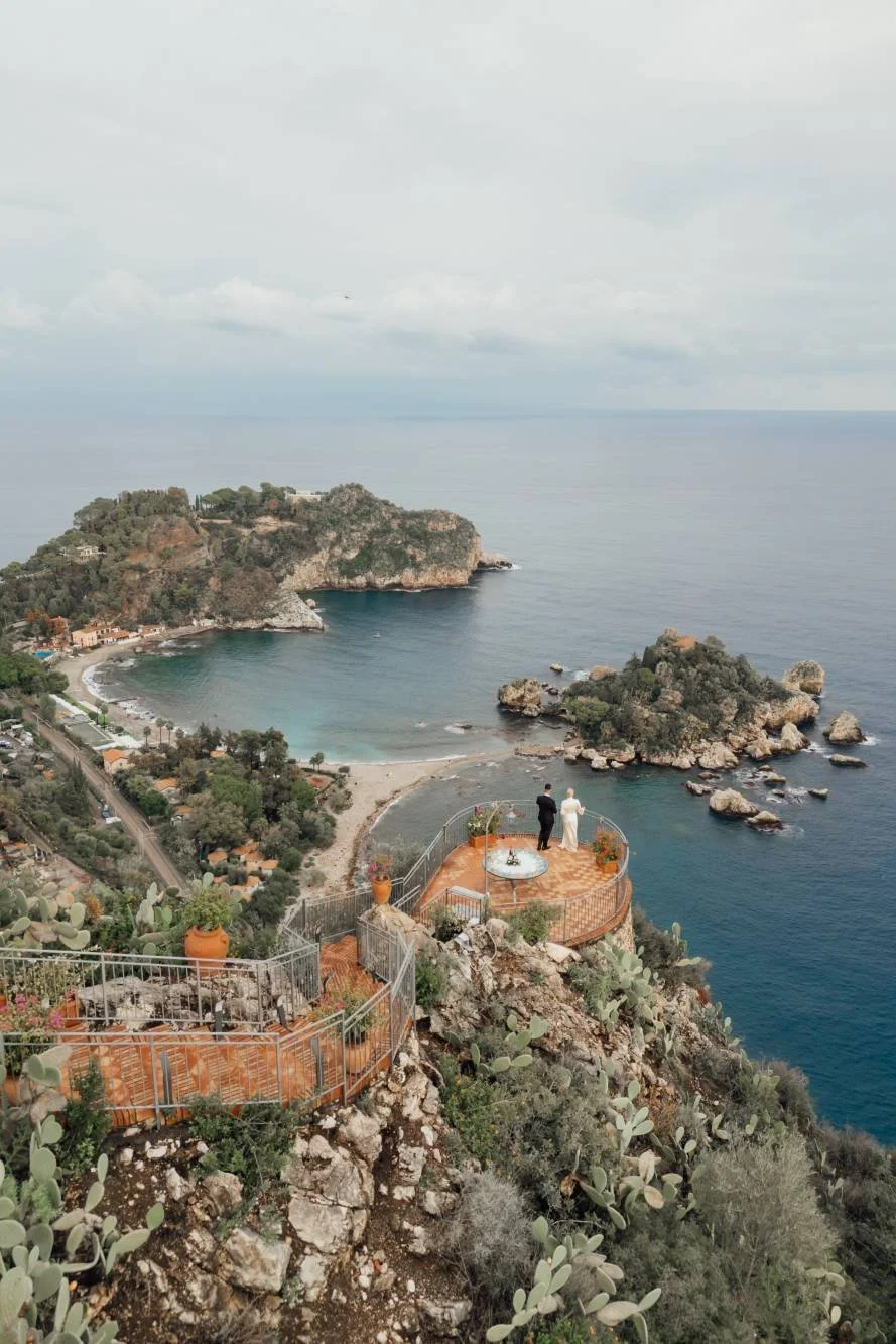 A coastal cliff with a wedding ceremony taking place on a terrace overlooking the sea, with two people standing at the altar and small boats on the water.