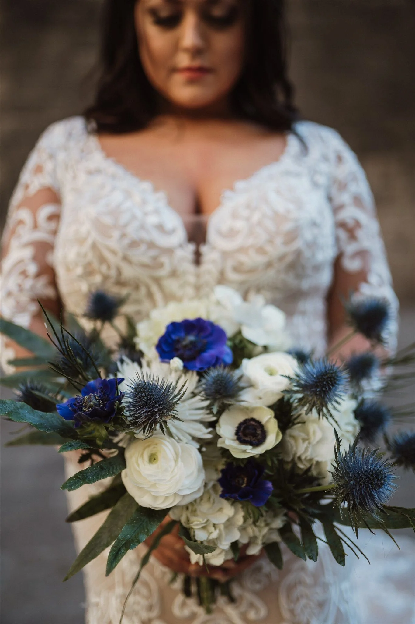 A woman in a white lace dress holding a bouquet of white, purple, and blue flowers, with thistle and greenery, standing on a decorative surface.