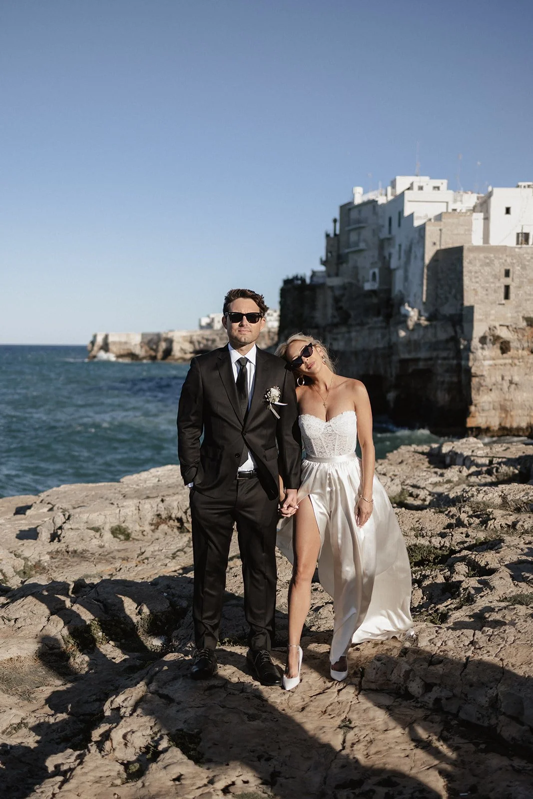 A man in a black suit and a woman in a white wedding dress stand on a rocky coastline holding hands, with buildings on a cliff and the ocean behind them.