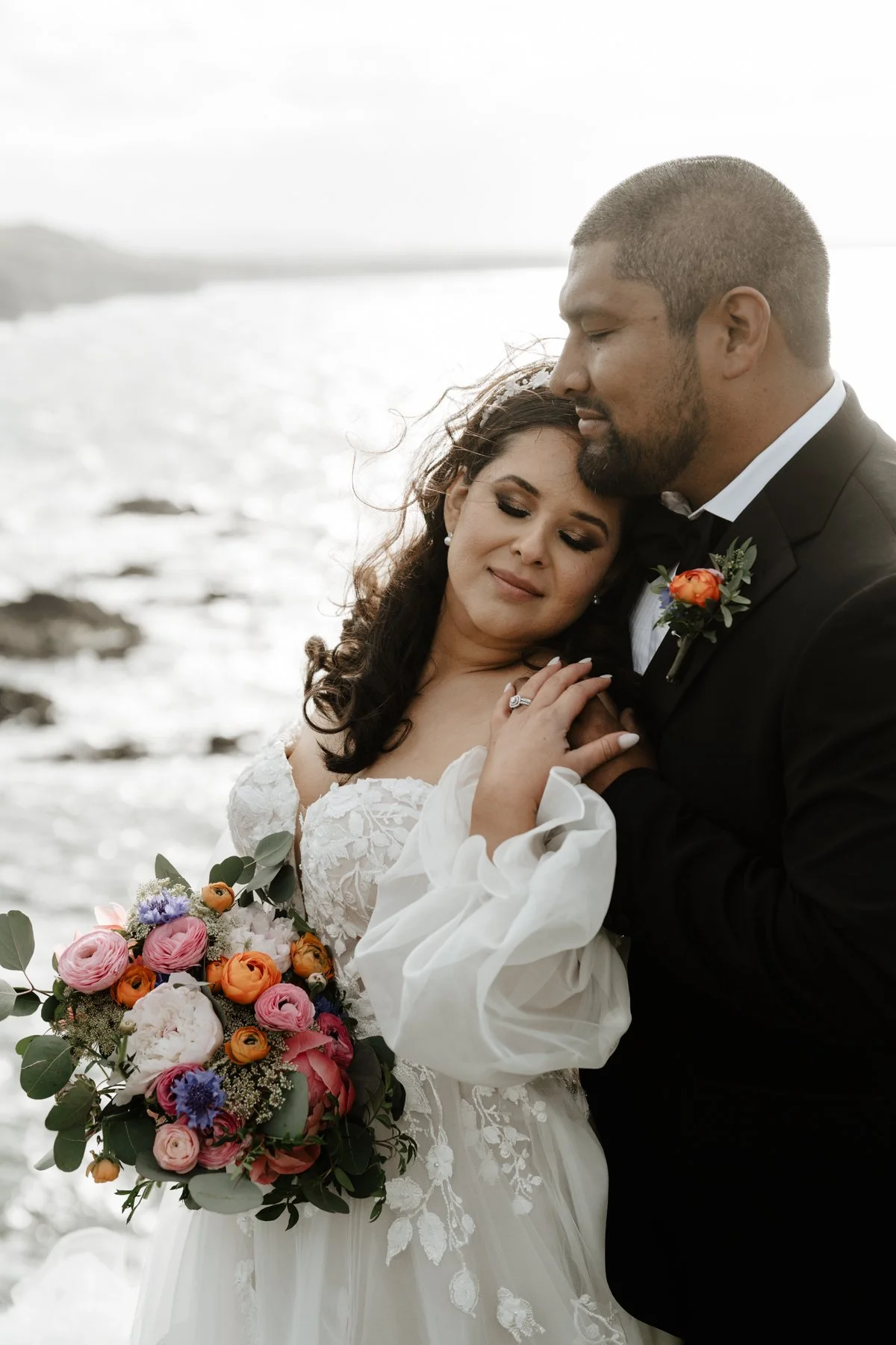 A bride and groom embrace during their wedding at a beach, with the bride holding a colorful bouquet of flowers.