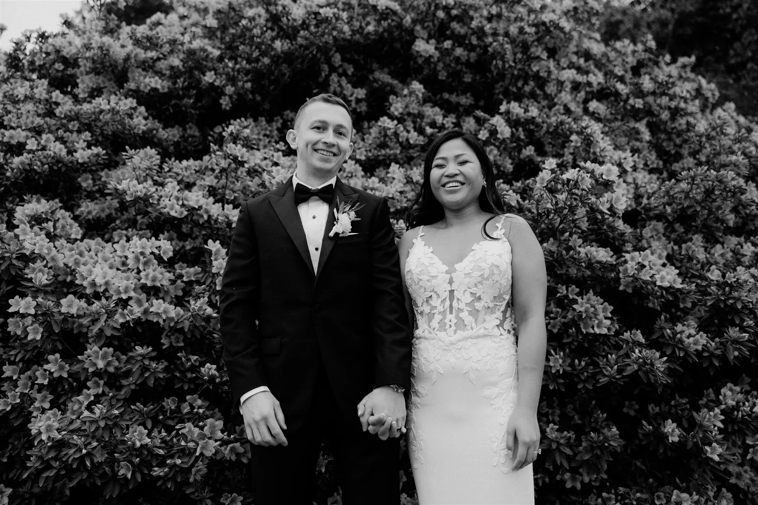 A black-and-white photo of a smiling couple holding hands in front of a large bush with flowers. The man is wearing a tuxedo with a bow tie and boutonniere, and the woman is wearing a wedding dress with floral lace details.