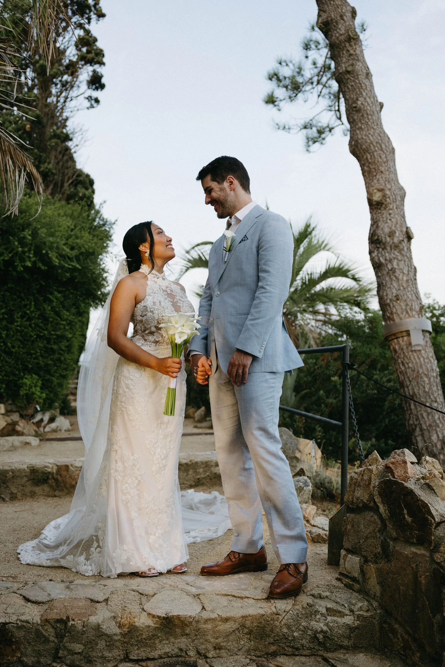 A bride and groom are holding hands and looking at each other during their outdoor wedding ceremony. The bride is wearing a lace wedding dress and holding a bouquet of calla lilies. The groom is dressed in a light blue suit with a white shirt and bro