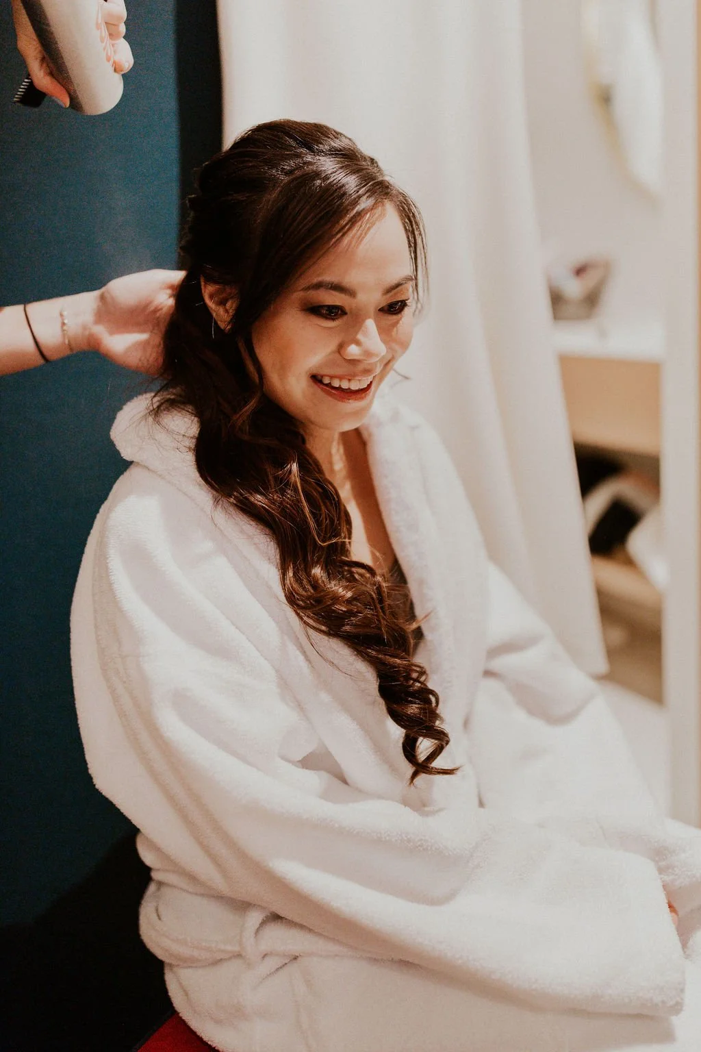Woman with long dark wavy hair getting ready, wearing a white bathrobe, smiling, in a room with plain background and a mirror.