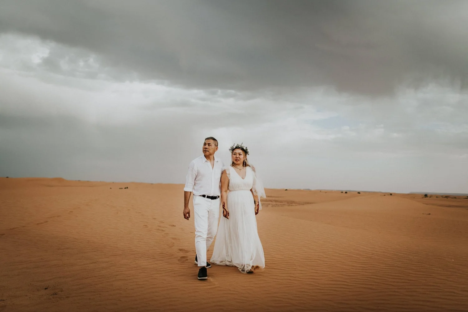A couple stands holding hands in a desert landscape with dark, cloudy skies overhead. The man, on the left, wears a white shirt and white pants, while the woman, on the right, is dressed in a white gown with lace details and a floral crown on her hea