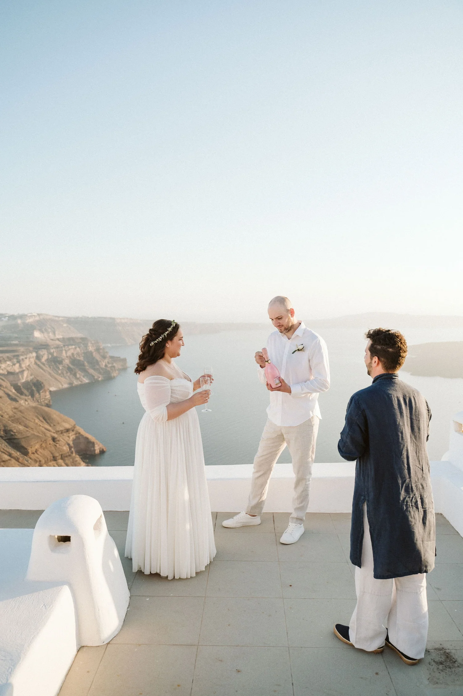A couple is getting married on a terrace overlooking the ocean and cliffs, with a man and woman officiant or witness present.