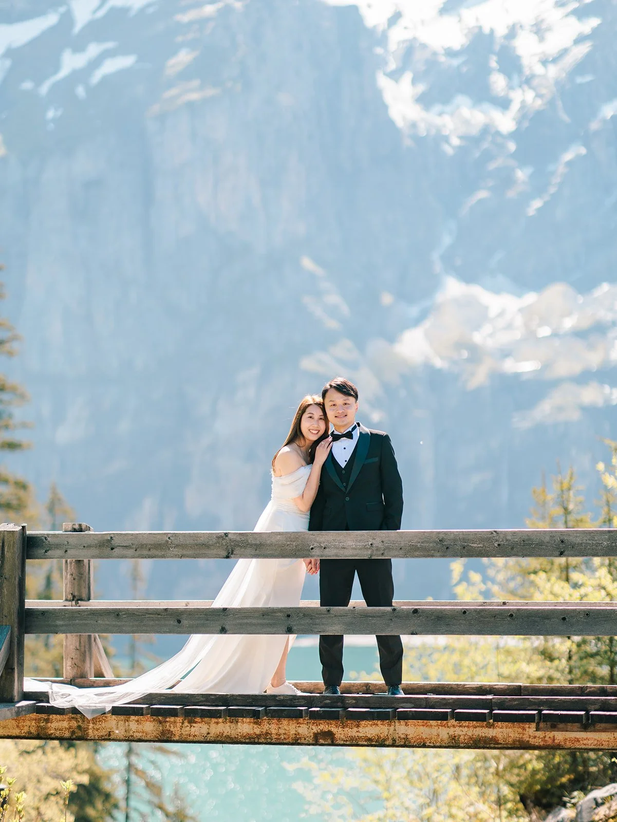 A bride and groom standing close together on a wooden bridge with a mountain and forest background. The bride is wearing a white off-shoulder wedding dress, and the groom is in a black tuxedo. They are smiling and smiling at the camera.
