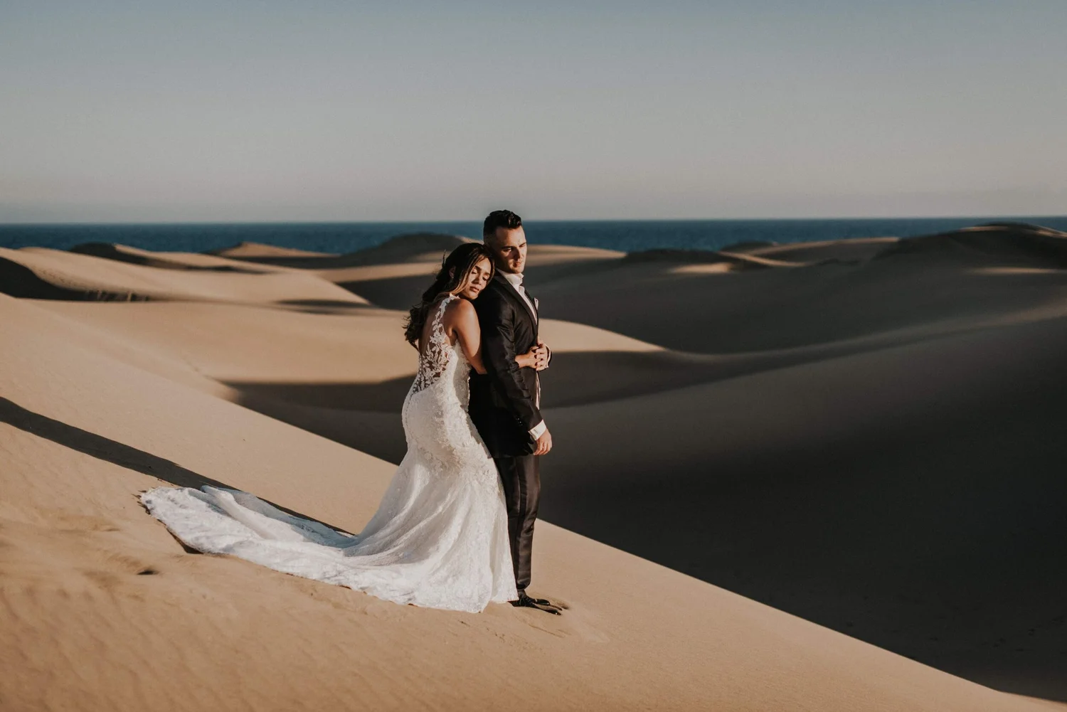 A bride and groom standing on sand dunes at the beach during sunset, with a wedding dress flowing behind the bride and the groom dressed in a black suit.