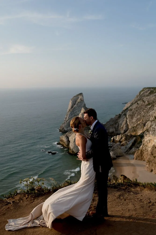 A bride and groom kissing on a cliffside near the ocean with large rocks and a sandy beach in the background.