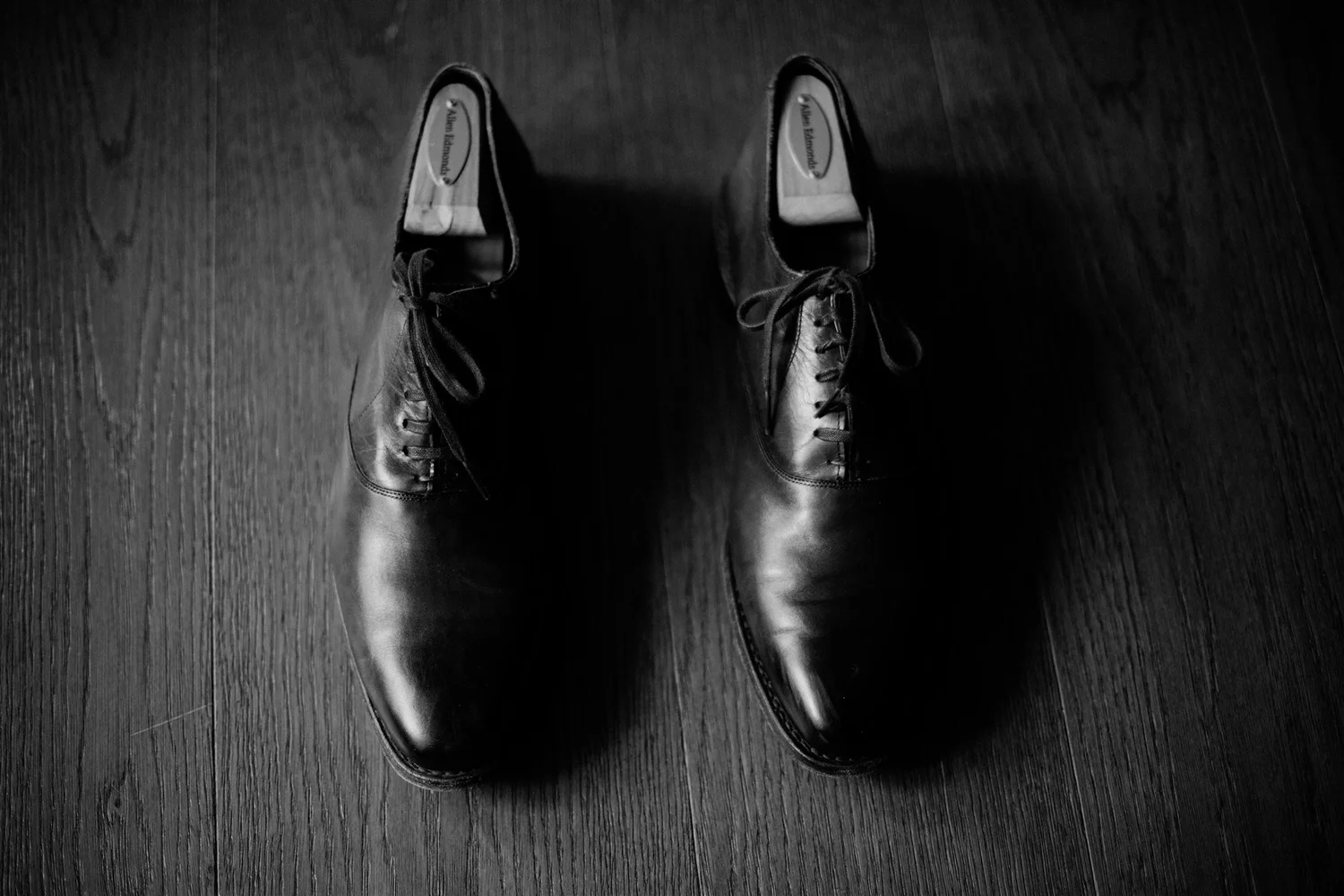 Black leather dress shoes worn on a person standing on a dark wooden floor.