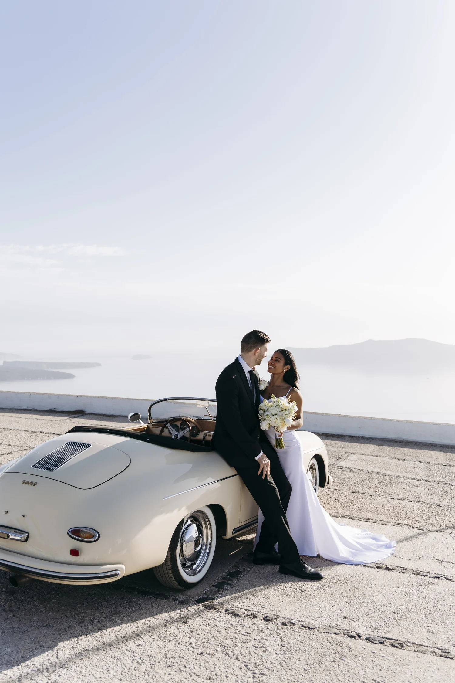 A bride and groom stand close together in front of a vintage white convertible car, with a scenic ocean view in the background, sharing an intimate moment.