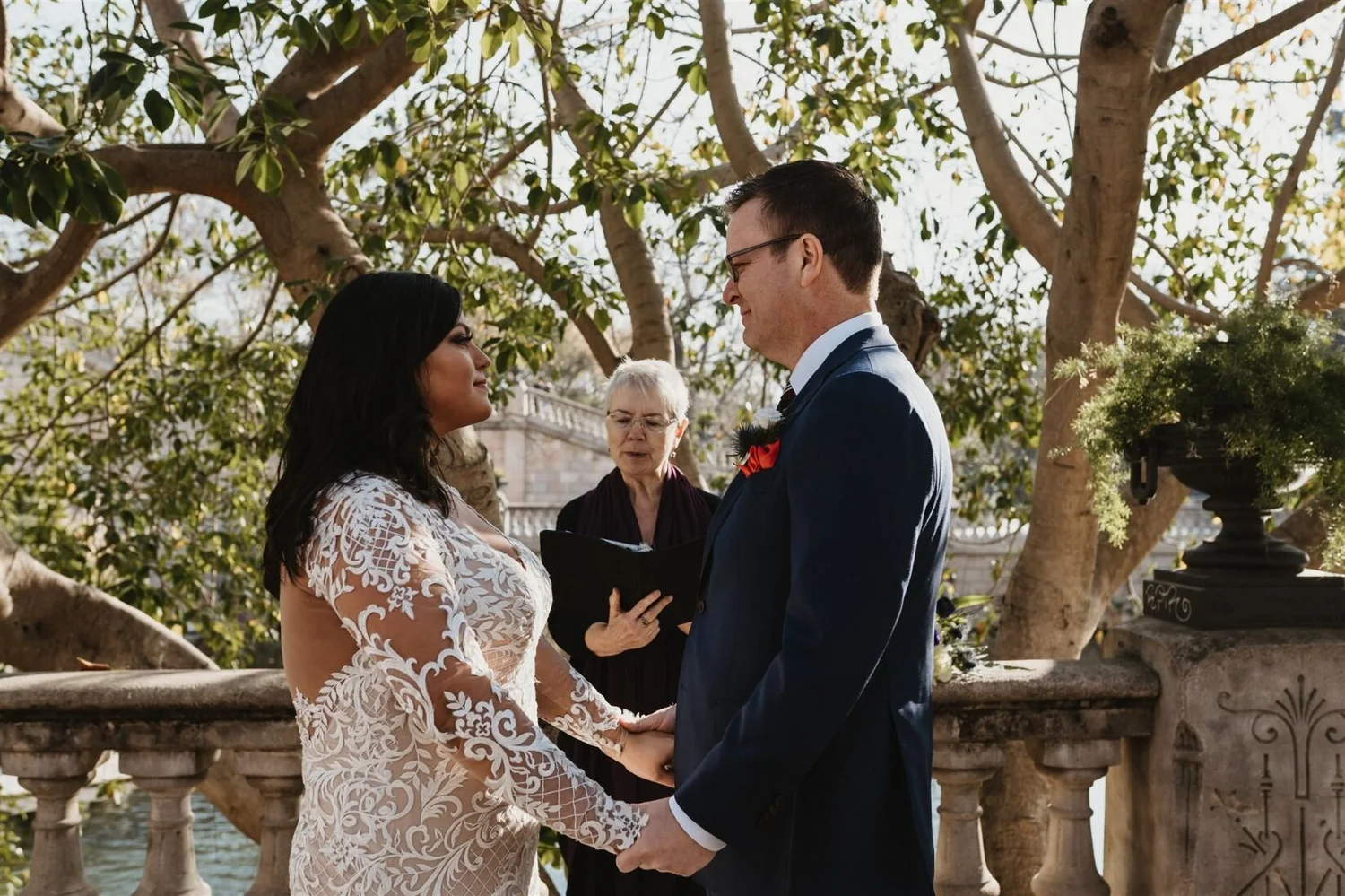 A couple gets married outdoors, holding hands and smiling at each other, with a woman officiant standing behind them, holding a book, in front of a tree with green leaves and a stone railing.