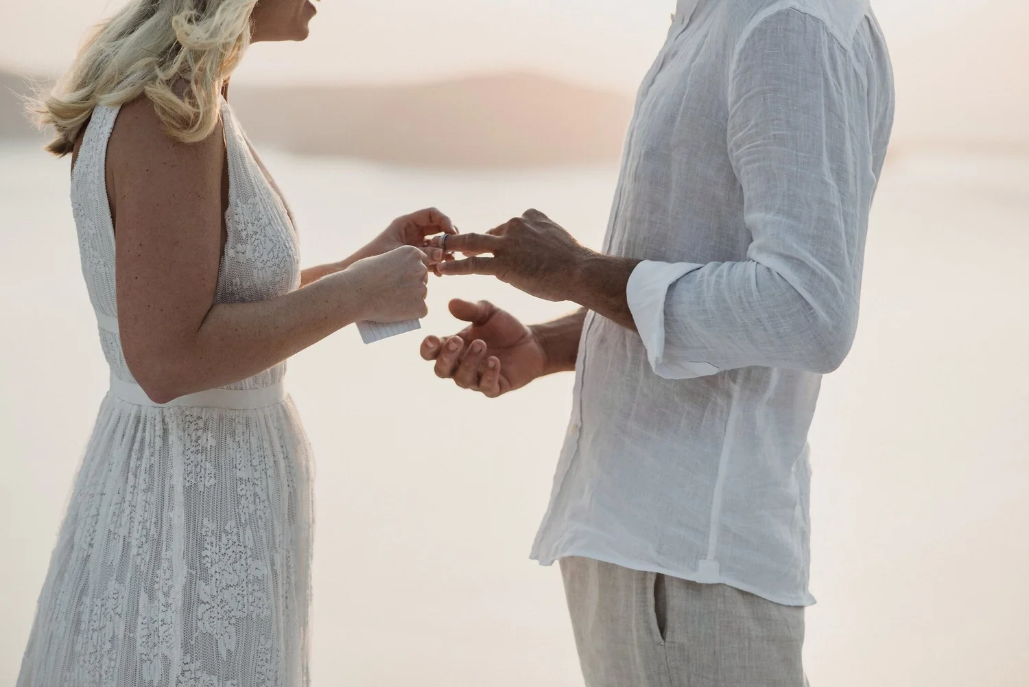 A couple exchanging rings outdoors near a body of water during sunset or sunrise.