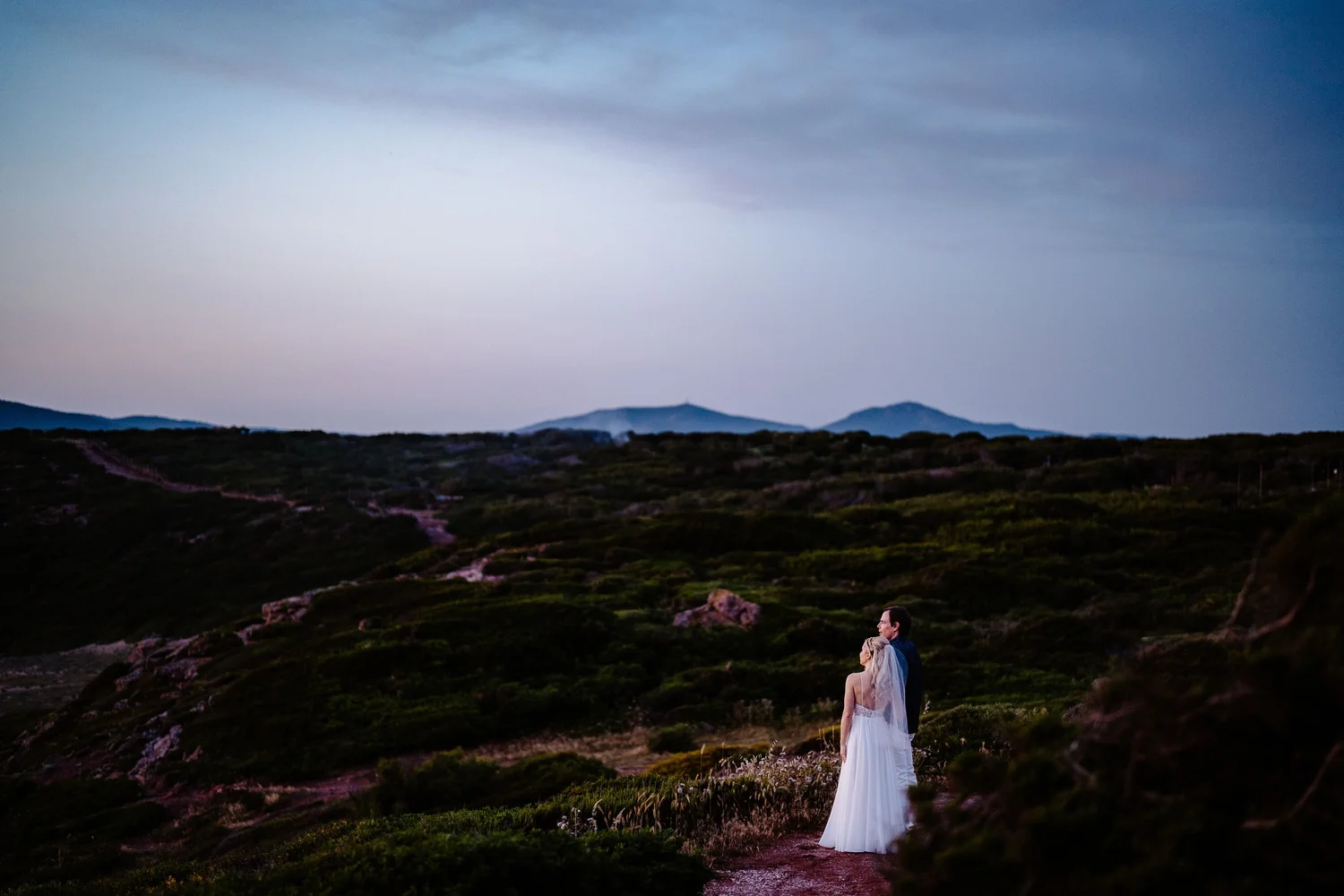 A bride and groom standing on a trail in a hilly landscape during twilight, with mountains in the background and clouds in the sky.