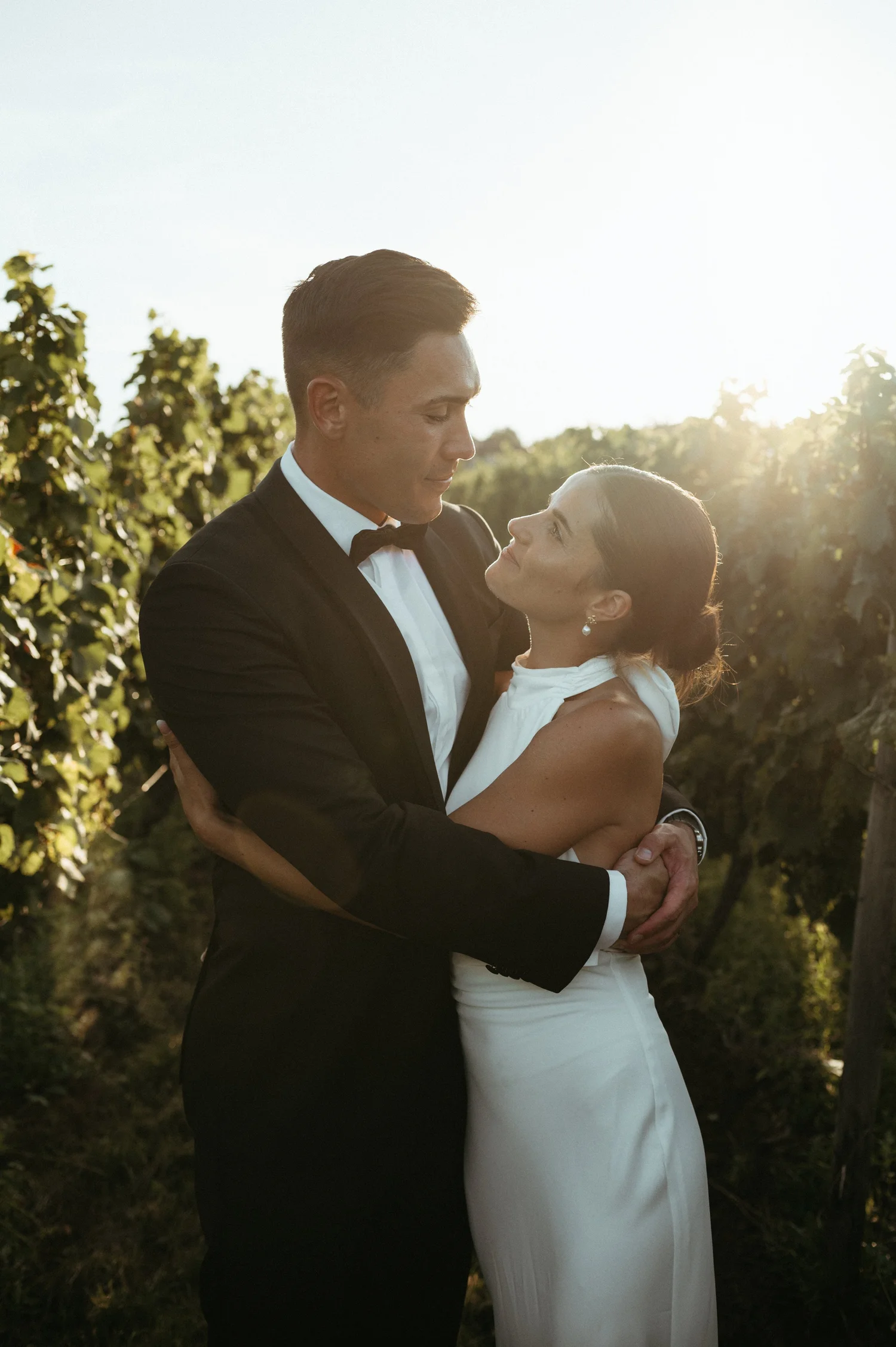 A man and woman dressed in formal attire, embracing outdoors in a vineyard at sunset.