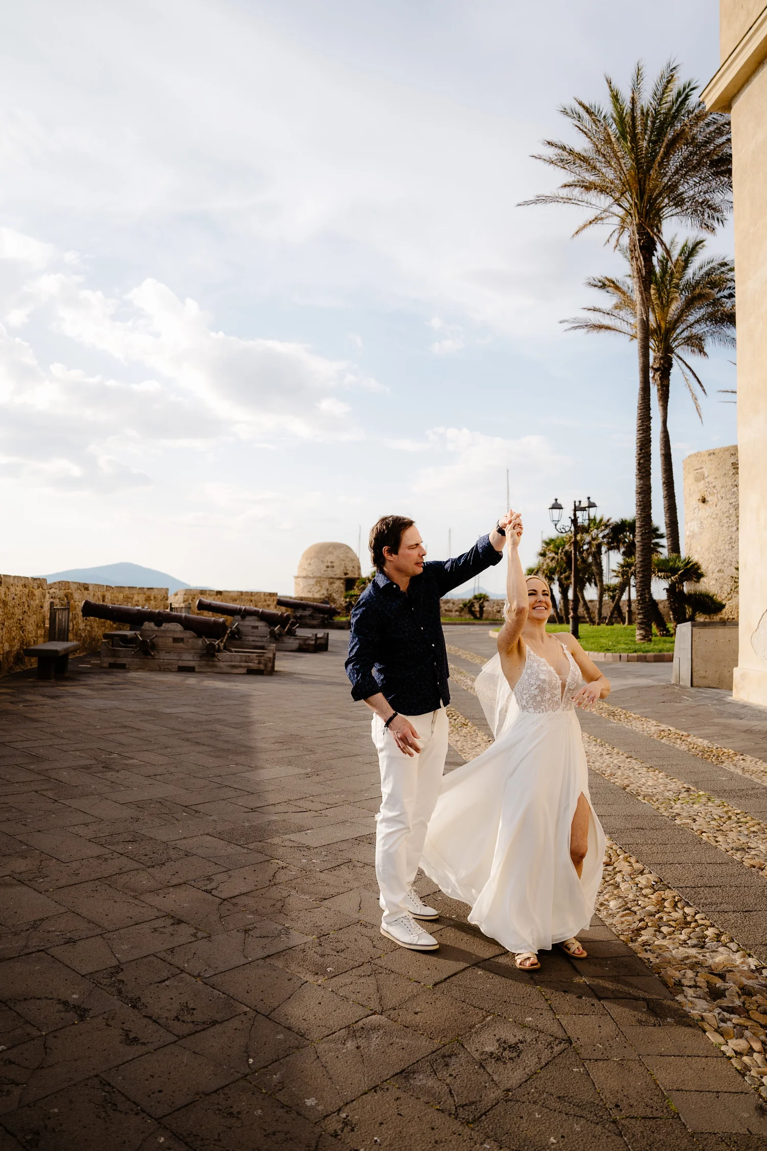 A newly married couple dancing outdoors during daylight, with palm trees and historic cannons in the background.