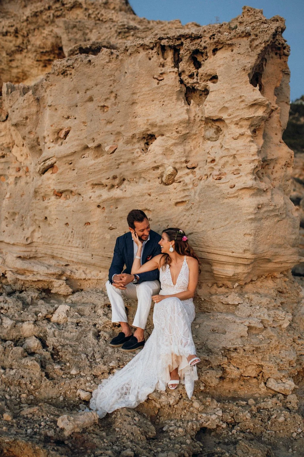 A couple dressed in wedding attire sitting close together on rocky ground in front of a large rock formation, smiling and looking at each other.