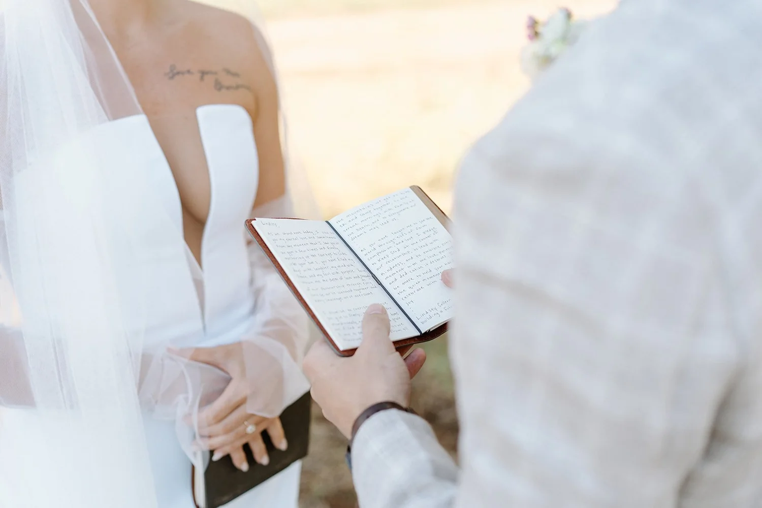 A person holding an open notebook with handwritten text, standing outdoors on a sunny day. Another person, dressed in white, is visible from behind, possibly a bride in a wedding dress, with a tattoo on their arm. The scene suggests a wedding or roma