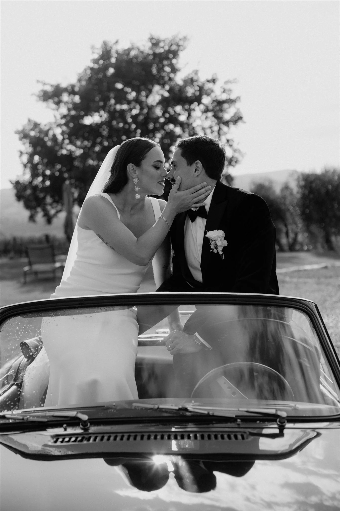 Black and white photo of a bride and groom in a car, with the bride touching the groom's face, both smiling, outdoors with a large tree in the background.