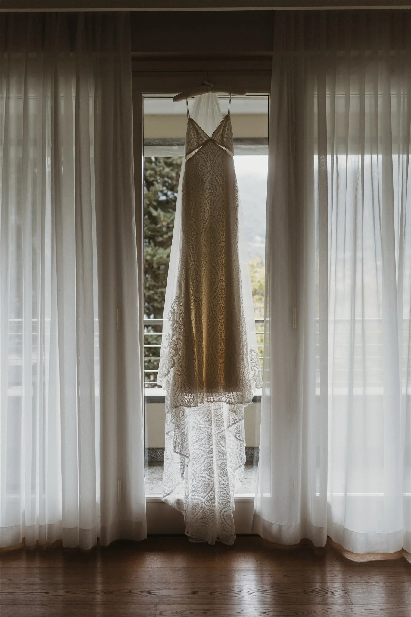 A wedding dress hanging on a curtain rod in front of a large window with sheer curtains, with trees and sky visible outside.