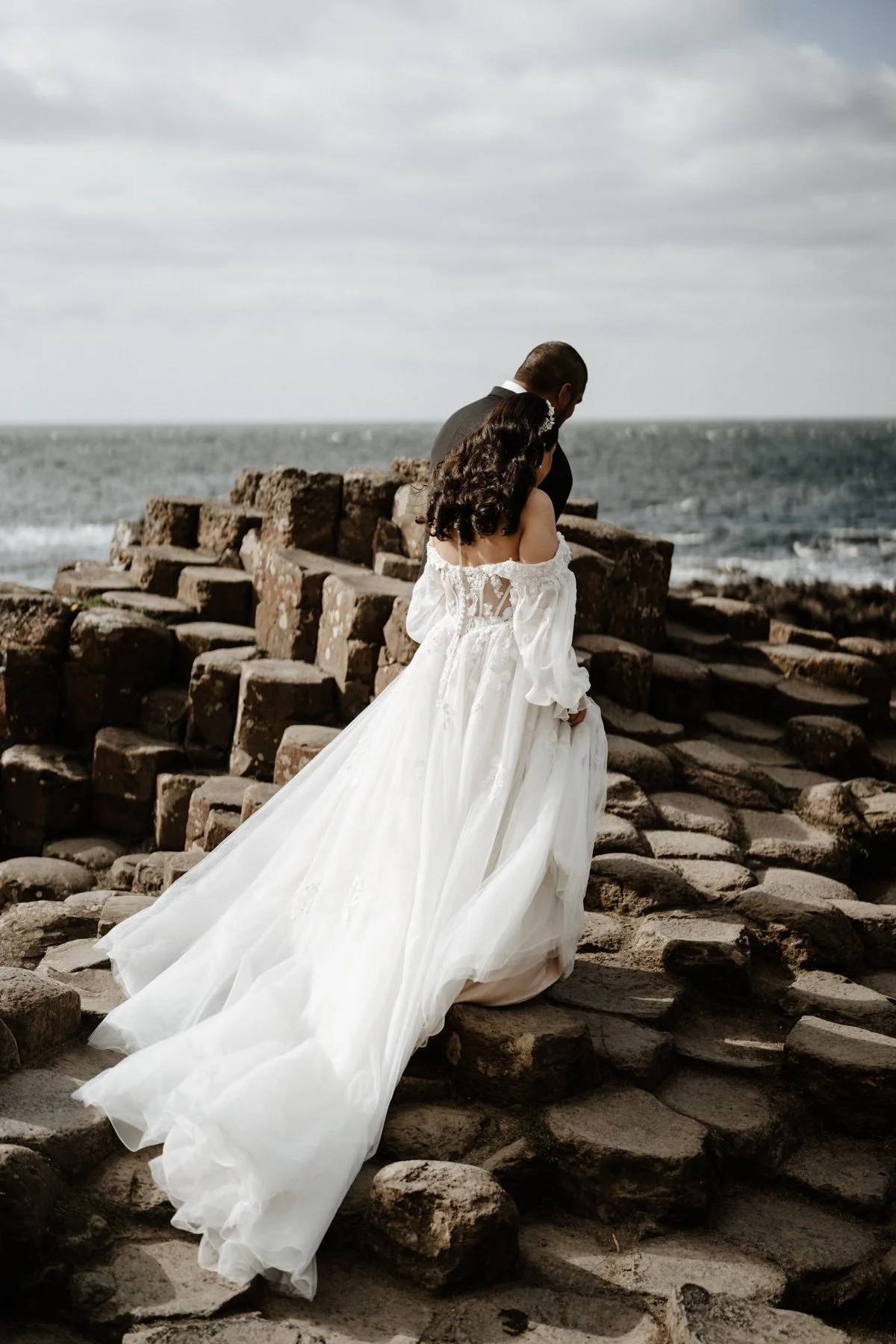 A bride in a white wedding dress walking on rocky steps near the ocean with a man in a black suit, both facing away from the camera, with a cloudy sky overhead.