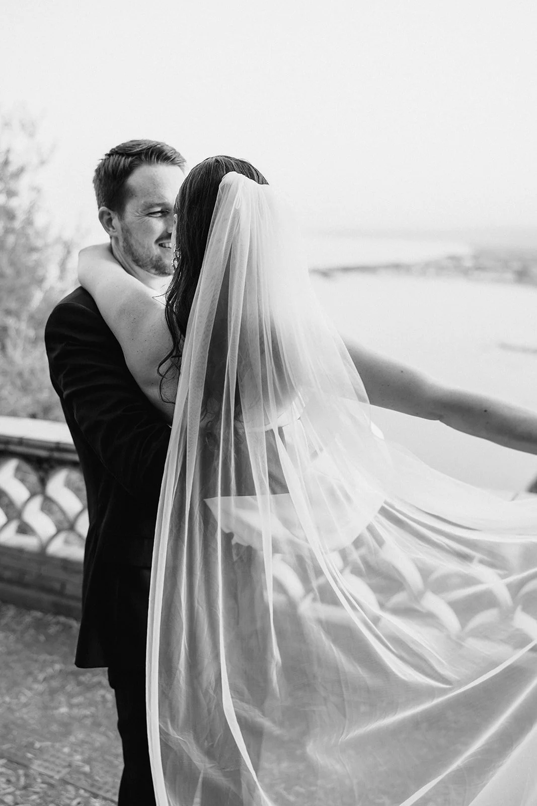 A black and white photo of a bride and groom embracing outdoors, with the bride's veil flowing and the groom smiling.