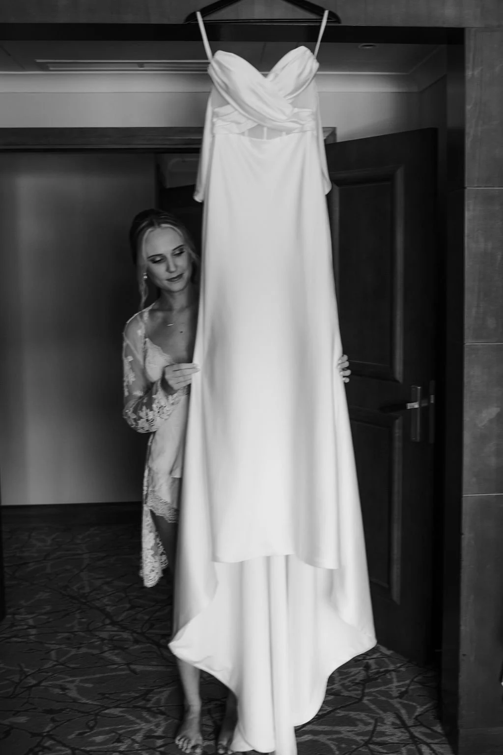 A woman in a lace robe standing behind a wedding dress on a hanger, looking at the dress, in a room with wooden walls and a patterned carpet.