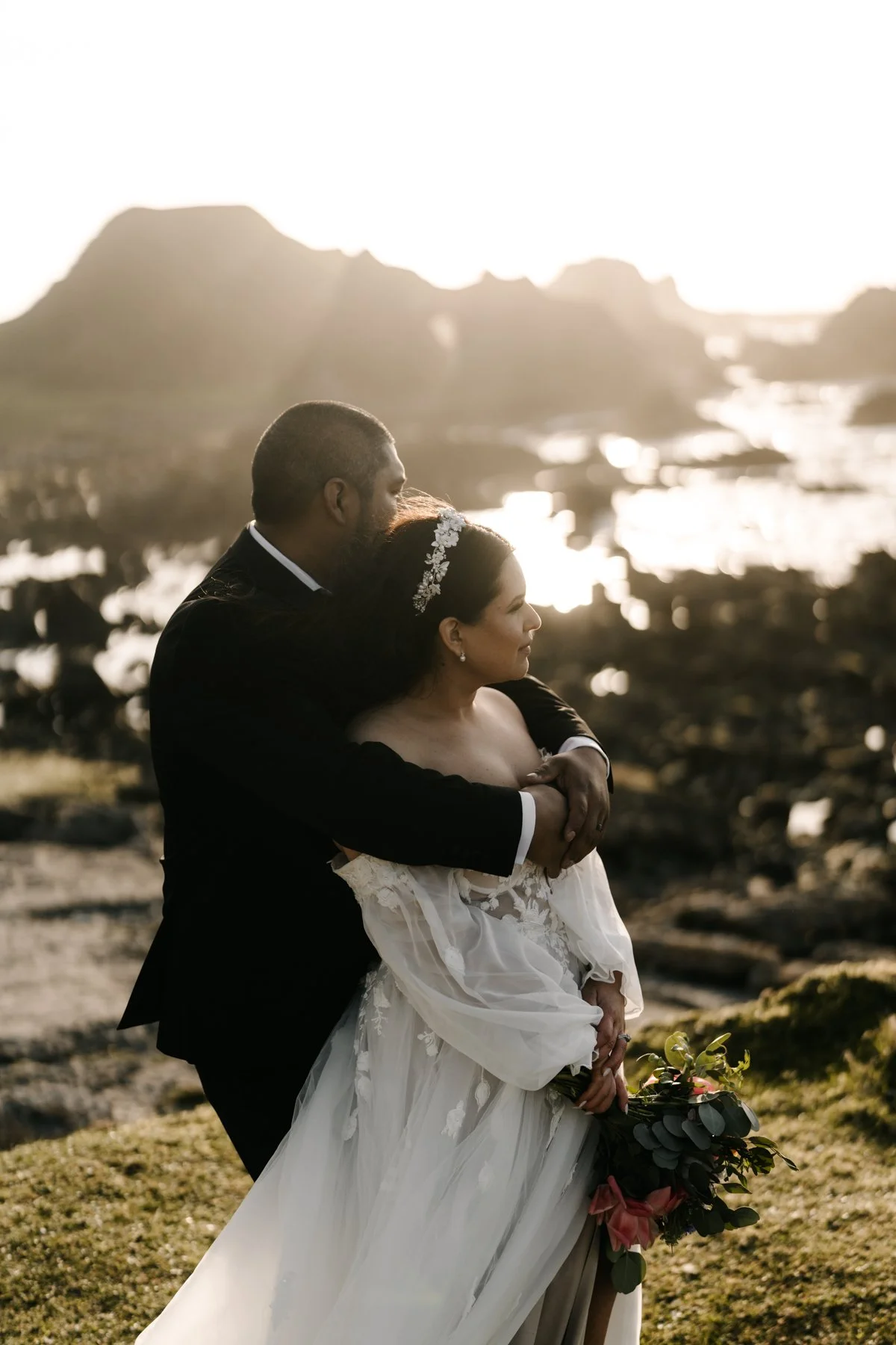 A couple dressed in wedding attire embracing outdoors during sunset, with a rocky landscape and water in the background.