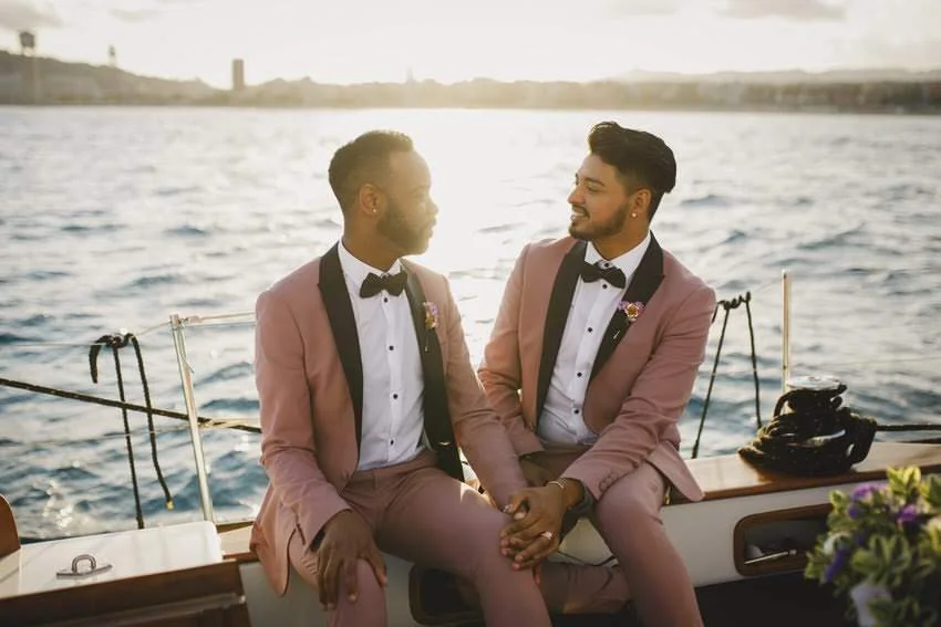 Two men in pink tuxedos with black lapels and bow ties sitting on a boat, holding hands, with water and a sunset in the background.