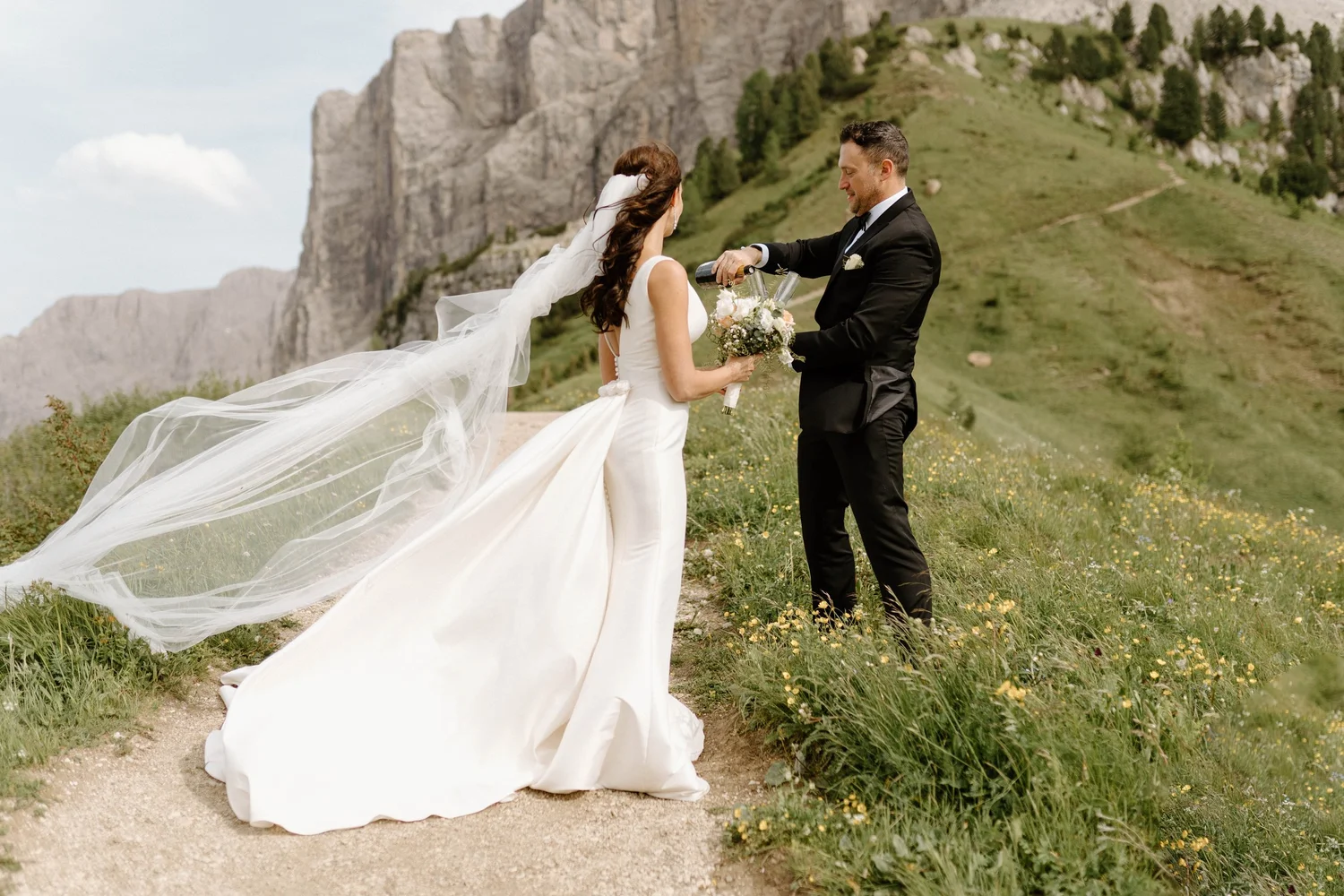 A bride and groom on a grassy trail in a mountainous area, with the bride holding flowers and the groom pouring champagne, celebrating after wedding.