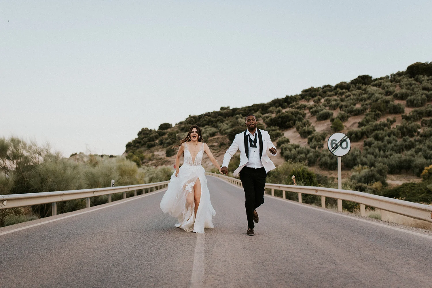 A bride and groom running happily hand in hand on an empty road in a scenic rural area.