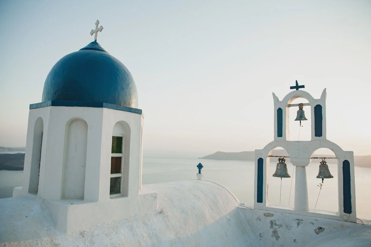 White chapel with blue dome and cross on top, white wall with small arched windows, and a bell tower with bells and cross against a pale sky.