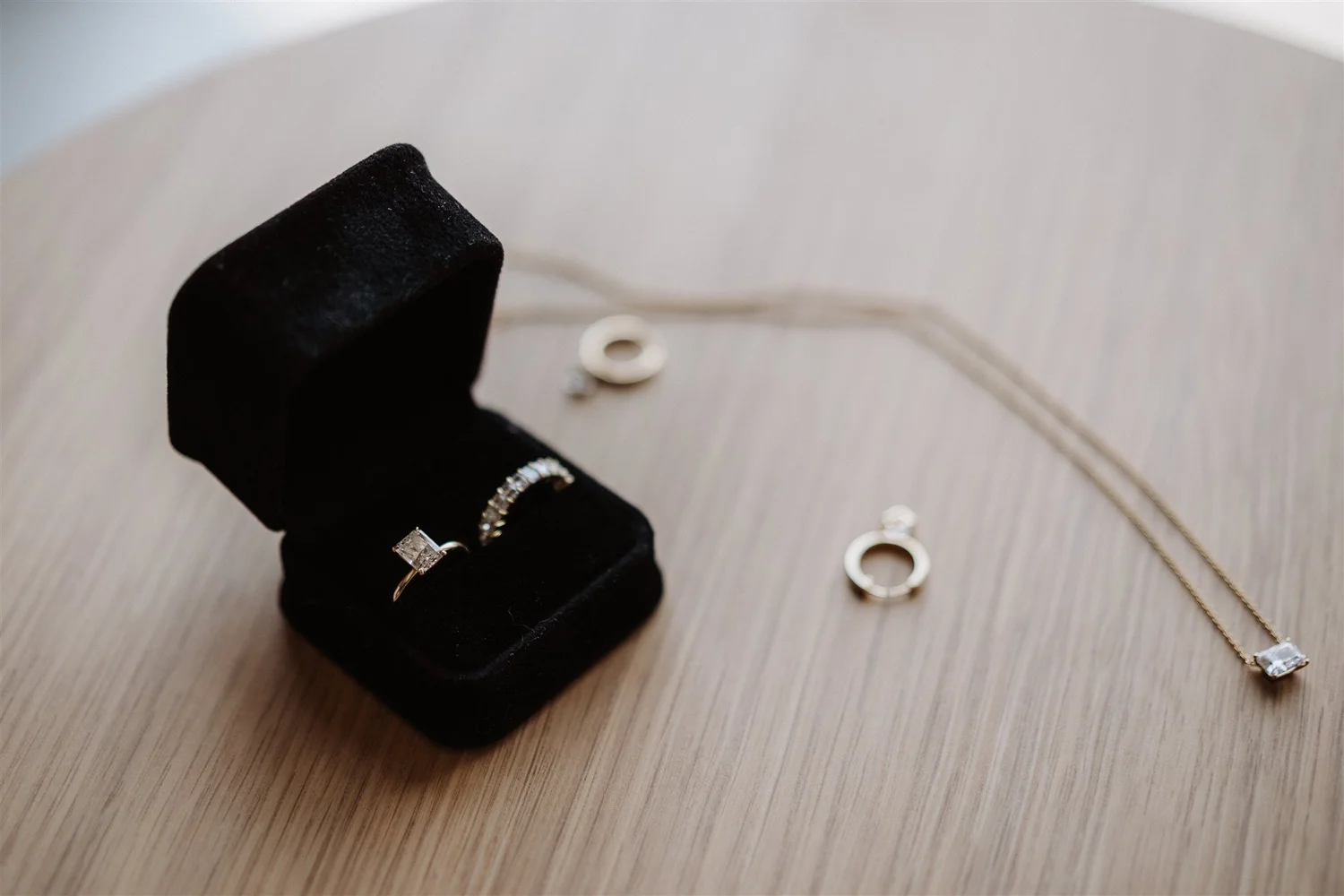 Close-up of a black jewelry stand displaying a silver ring with a square gemstone, in front of silver hoop earrings and a necklace, all on a wooden surface.