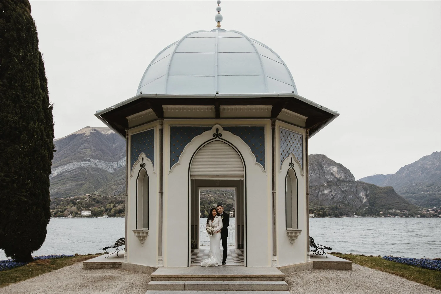 A bride and groom standing under a small ornate pavilion overlooking a lake with mountains in the background.