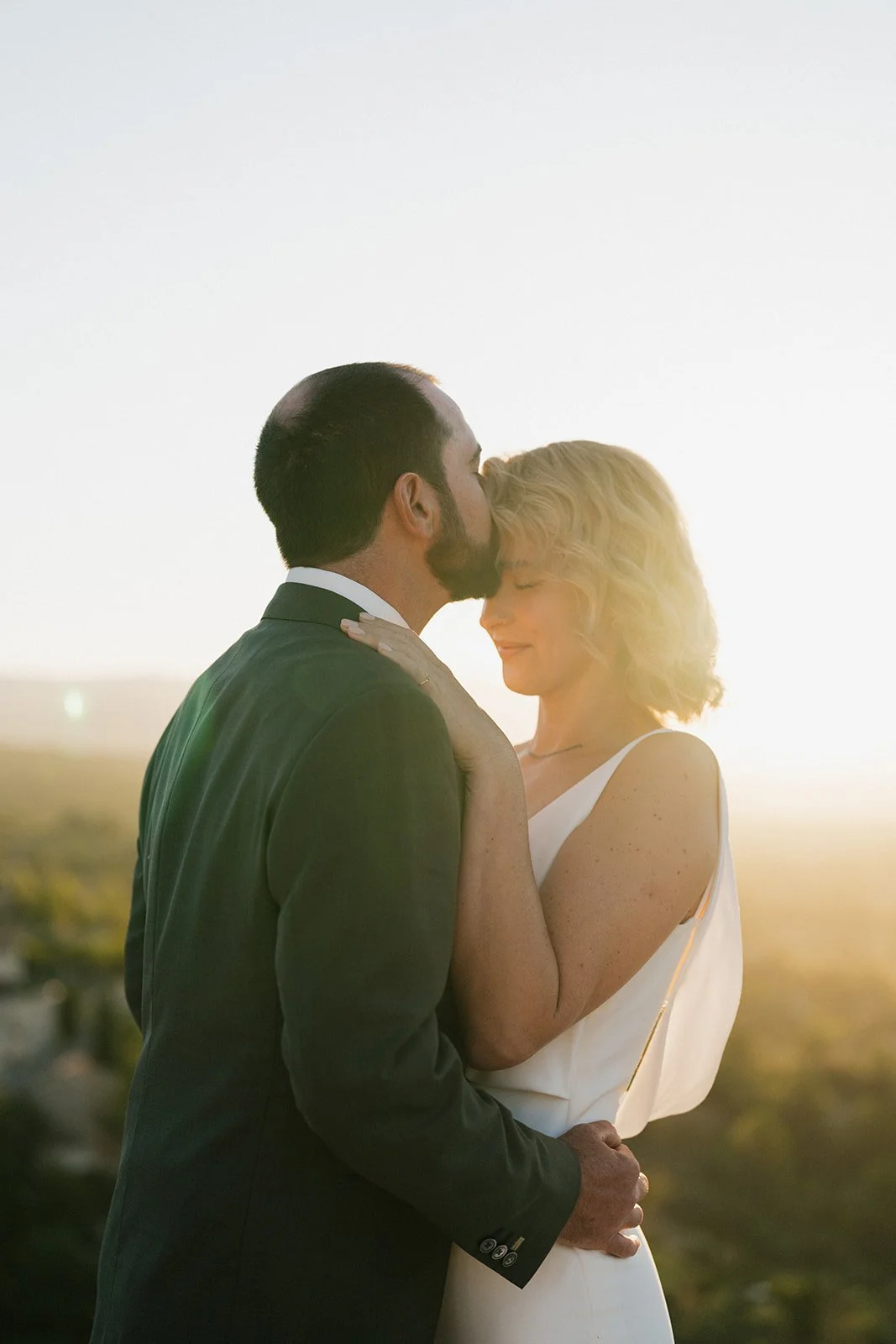 A couple dressed formally, sharing an intimate moment at sunset outdoors, with the man kissing the woman's forehead and the woman smiling with her eyes closed.