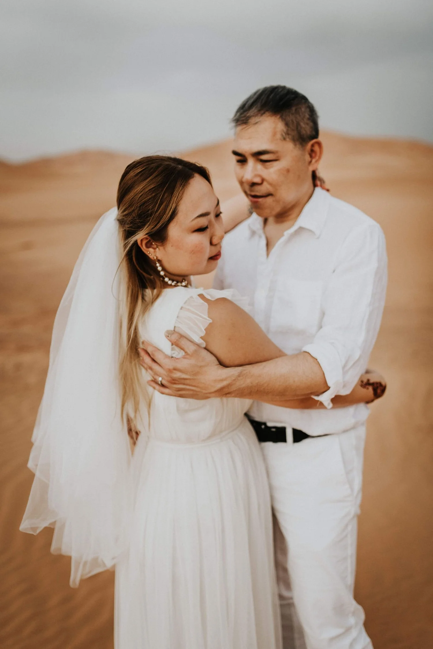 A couple dressed in white, embracing in a desert landscape with sand dunes in the background.