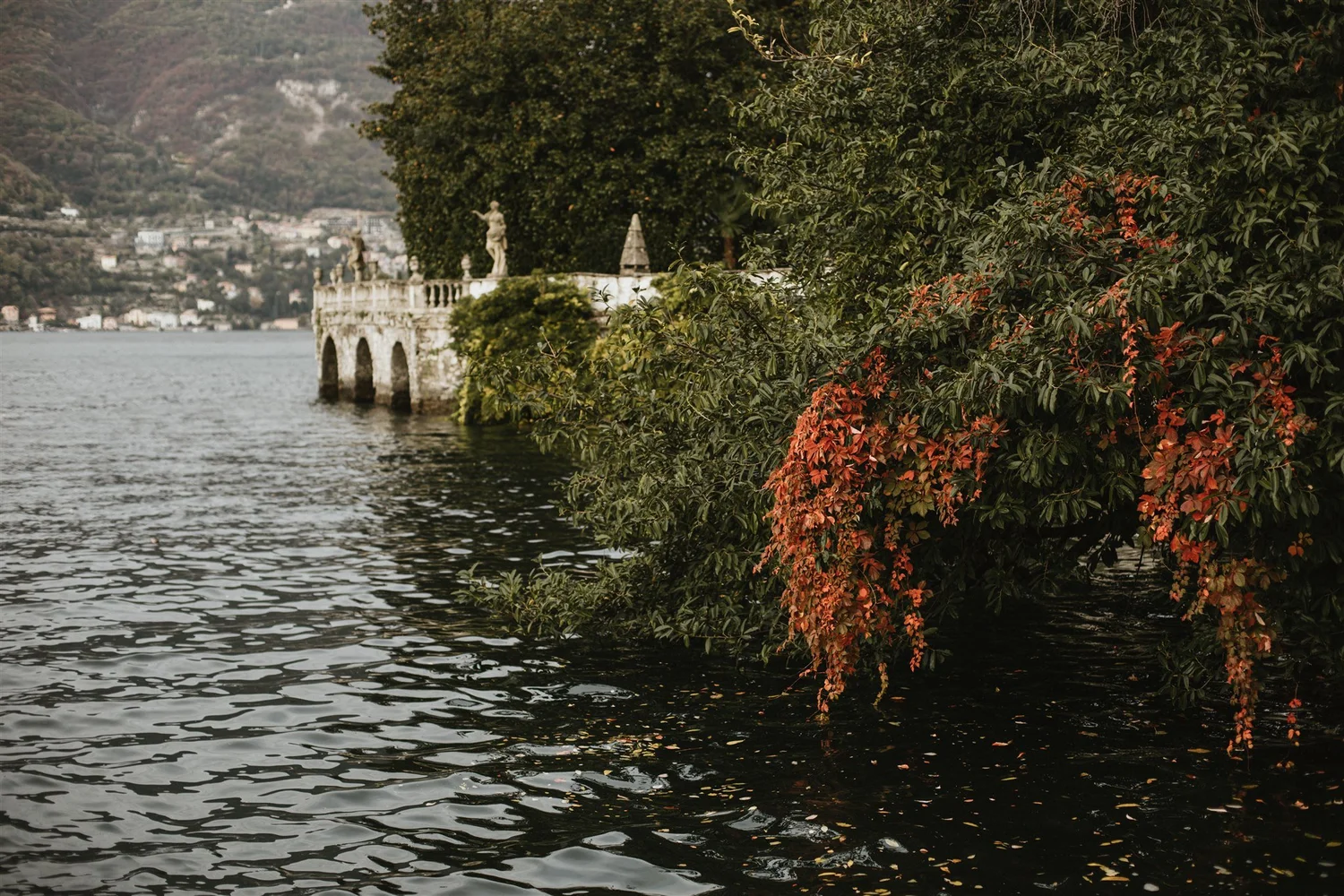 A lakeside scene with lush green trees, some with red-tinged leaves, partially covering a historical stone structure with statues on a terrace. The lake water is visible in the foreground, and a town with buildings and hills is in the background.