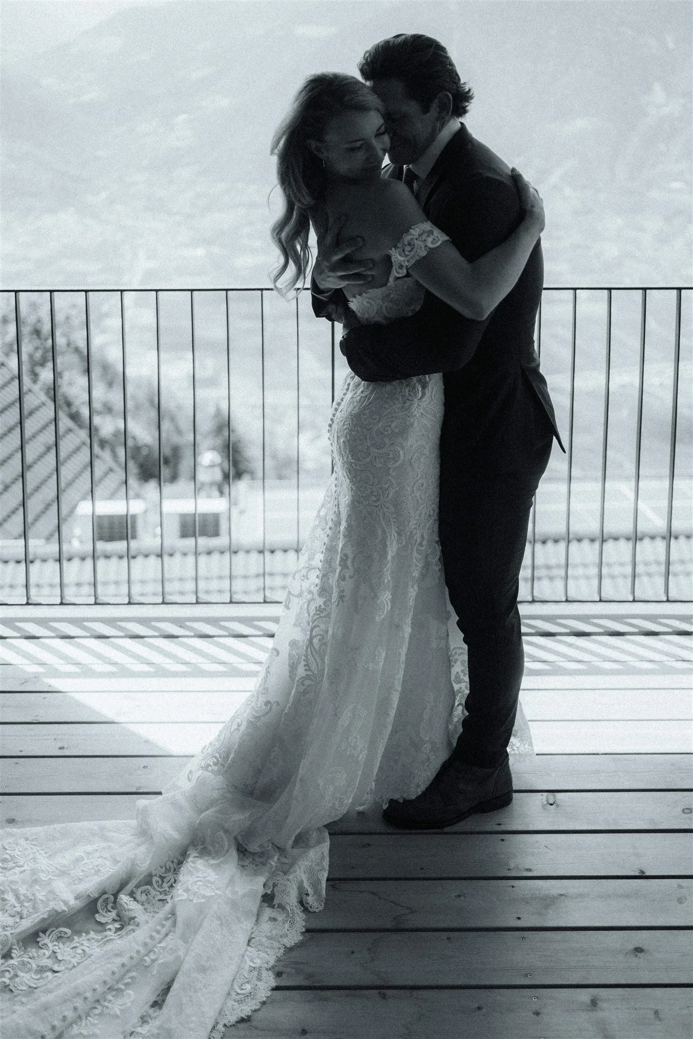 A bride and groom embracing on a deck, with the bride wearing a detailed lace wedding gown and the groom in a dark suit. They are standing close to a railing with a cloudy sky in the background.