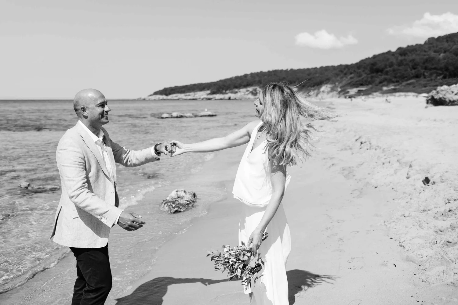 A man in a light-colored blazer and a woman in a white dress exchanging vows on a beach, with hills and a cloudy sky in the background.