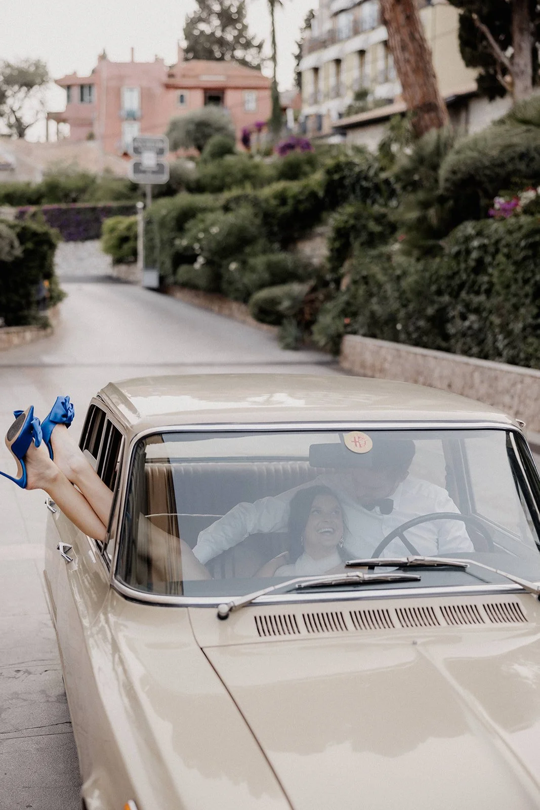 A woman with high heels feet sticking out of the car window, smiling and looking at a man driving, inside a vintage beige car on a city street with greenery and buildings in the background.