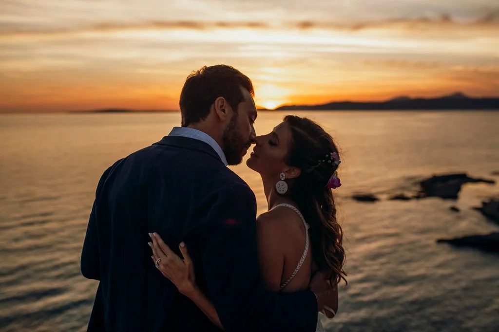 A couple kissing at sunset by the water, with rocky shoreline in the background.