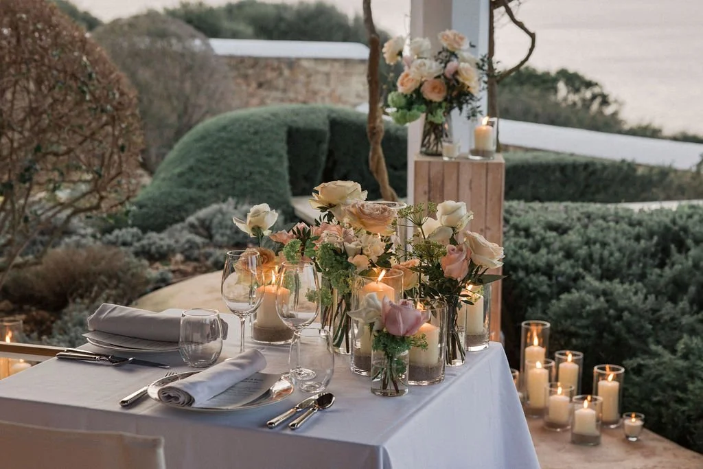 Elegantly set outdoor dining table with white tablecloth, floral centerpieces, candles, and glassware, overlooking a garden with bushes and trees.