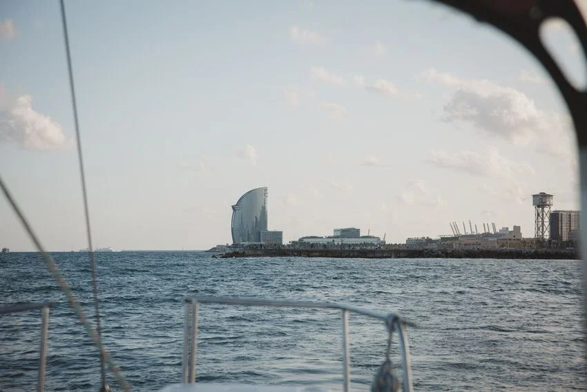 View of the ocean with a city skyline including a distinctive sail-shaped building, likely the W Barcelona hotel, as seen from a boat.